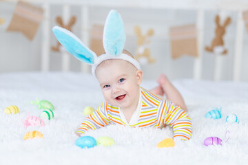 Wall Mural - baby boy with rabbit ears on his head lying on the bed with Easter eggs, cute funny smiling little baby. The concept of Easter.