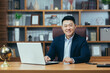© Liubomir - Portrait of a successful Asian businessman, man working in the office sitting at the table, looking at the camera and smiling, happy banker with laptop