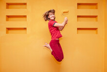Young Woman In Red Closing With A Brush Jumping Happily In Front Of Yellow Walls While Painting.