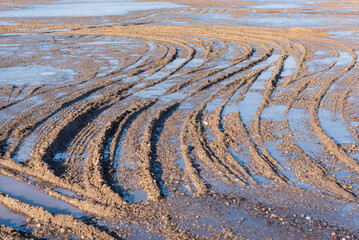 Country road with melting ice and puddles, sunny spring day. Traces of cars on the frozen ground.