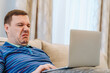 Annoyed millennial man with disgust confused looking at laptop screen sitting on sofa at living room. cringe guy looking at computer with squeamishness, reacting on watching disgusting video at home