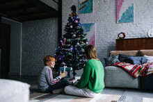Teens Sitting Near Decorated Conifer Tree And Give Gift