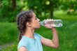 Brunette caucasian girl with braided hair is drinking water from a plastic bottle in green summer park