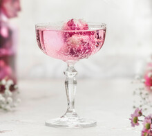 Close Up Of Sparkling Pink Drink With Rose Petals In Champagne Glass At White Table With Blurred Background. Aperitif With Elegant Decoration For Celebration. Front View.