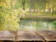 Empty Wooden Aged Table Surface Against The Background Of The Lake. A Flowering Tree In The Background And The Paths Of The Park In Defocus. Spring Flowering