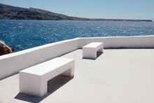 Two White Benches On The Coast Of Santorini And The Sea With Blue Sky And Mountains In The Background. Public Place On Santorini Island, Oia, Greece. Beautiful Seascape View With Space For Text