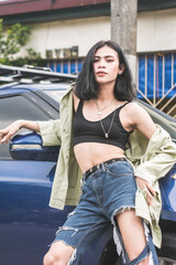 A confident and fashionable asian trans woman posing in front of a blue car. Wearing ripped jeans and a black crop top. Outdoor modelling scene.