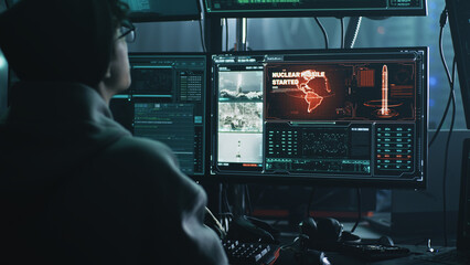 Young male hacker in hat and glasses using computers to hack nuclear warhead and start war while sitting at table in dark room