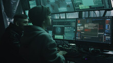Two Hackers In Casual Clothes Cracking Classified Files On Computer And Infecting Documents With A Virus In A Dark Room With Monitors
