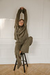 © Look! - Full length young caucasian woman sits on stool stretching arms up against white background with space for text. Tanned lady wears glasses, grey warm suit with eyes closed. Weekend enjoyment concept