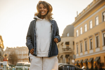 Wall Mural - Funny fair-skinned young girl jumps on spot, smiling broadly to her teeth in city center. Tanned blonde with tousled hair is wearing white tracksuit and denim jacket. Holiday life concept