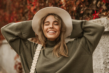 Beautiful young caucasian girl closing her eyes holding hat behind head in fresh air. Blonde is smiling broadly with her teeth, happy in park on autumn day. People sincere emotions lifestyle concept.