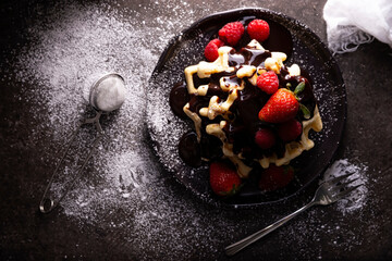 Wall Mural - Top view of homemade waffles with chocolate, icing sugar, strawberries and raspberries on dark dish. Messy look flat lay.