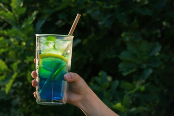 Wall Mural - Tall glass with a blue lagoon cocktail in a female hand against the backdrop of greenery in the yard. Selective focus