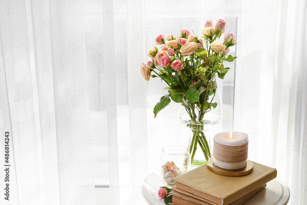 Vase with roses, books and burning candle on table in light room