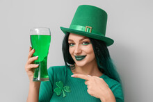 Young Woman In Green Wig Pointing At Glass Of Beer On Light Background. St. Patrick's Day Celebration