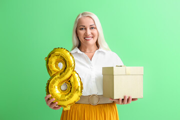 Poster - Smiling mature woman holding gift box and balloon on green background. International Women's Day celebration