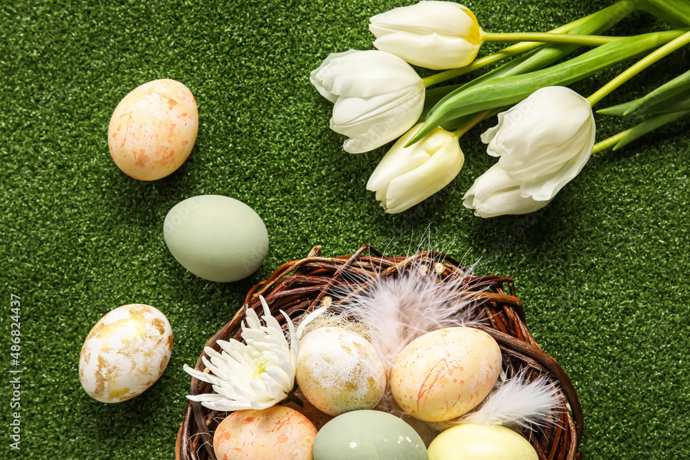 Nest with painted Easter eggs, feathers and flowers on green background