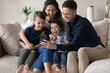 © fizkes - Happy young parents and excited kids watching funny video online content on mobile phone, making call, talking, using app, getting good news, feeling joy. Family using smartphone, relaxing on couch
