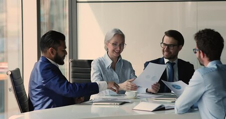 Canvas Print - Satisfied older age businesswoman young male investor reached agreement at round table discussion shake hands. Diverse partners handshaking after successful negotiations on business group conference