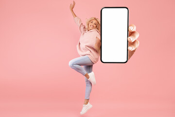 Positive woman jumping up and showing big blank cellphone screen advertising great app or website on pink background