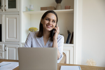 Wall Mural - Distracted from working on laptop happy young businesswoman listening pleasant news enjoying cellphone call conversation, discussing online project success with colleagues, multitasking in office.