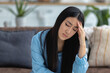 © kucherav - Asian young woman sitting on the sofa at home holding her head, headache, migraine, emotional stress