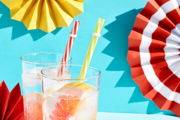 Wall Mural - Summer cocktail with fresh grapefruit, ice and slices of citrus fruit in highball glass on pastel blue surface with colorful paper fans as summertime party concept. Fresh healthy grapefruit beverage.