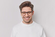 Photo of handsome cheerful man with dark hair smiles gladfully with closed eyes expresses positive emotions daydreams about something wears spectacles and casual jumper isolated on whie wall
