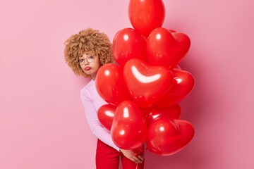 Wall Mural - Indoor shot of unhappy curly woman feels sad on Valentines Day frowns face has bad mood wears transparent eyeglasses jumper and trousers holds red inflated balloons isolated on pink background