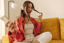 Fashionable European Girl In Good Mood Poses For Photo While Sitting On Couch. Beauty In Summer Outfit Relaxes While Listening To Favorite Song Through Headphones. Lifestyle Concept, Playful Mood.