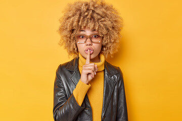 Wall Mural - Mysterious curly haired woman makes silence gesture presses index fingers to lips asks to be quiet makes taboo sign wears sweater and leather jacket isolated over yellow background. Do not speak