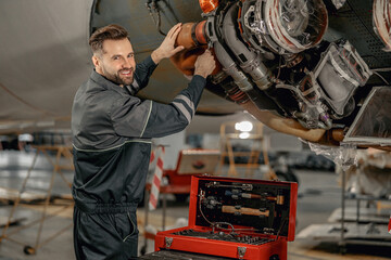 Cheerful man maintenance technician looking at camera and smiling while repairing airplane at repair station