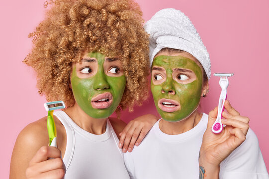 Embarrassed two young women hold razors going to do hygiene procedures shave legs apply green beauty mask made of cucumbers for skin moisturizing wear bath towel on head isolated over pink background