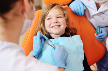 young dentists, a man and a woman, examine the teeth of a child's patient - a little pretty girl who is sitting in an orange dental chair.