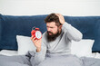 Perplexed guy scratching head holding alarm clock being in bed in morning, oversleep