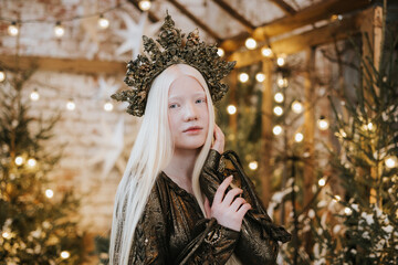 young albino woman with blue eyes and long white hair in beautiful green dress and crown stands in loft room decorated with wooden greenhouse and Christmas trees with twinkle lights, diverse people