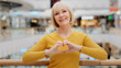 © Yuliia - Mature happy caucasian woman standing in public place posing sincerely smiling looking at camera making heart shape with fingers sign generosity and love mercy gesture kindness symbol friendliness