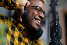 Close-up Of An African Podcaster Blogger Smiling While Broadcasting His Live Audio Podcast In The Studio Using A Microphone And Headphones. Male Radio Host With Glasses, Podcast Host Or Interviewer