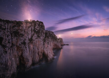 Lighthouse On The Mountain Peak At Starry Night In Summer. Beautiful Cliffs, Rocky Sea Coast, Bright Stars, Milky Way And Violet Sky With Clouds At Dusk. Lighthouse Of Cape Lefkada, Greece. Landscape