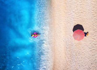 Poster - Aerial view of a woman swimming with pink swim ring in blue sea, sandy beach and red umbrella at sunset in summer. Tropical landscape with girl, clear water, waves. Top view. Lefkada island, Greece