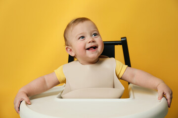 cute little baby wearing bib in highchair on yellow background