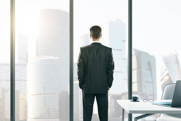 Sticker - Back view of thoughtful young businessman standing in modern office with workplace desktop and panoramic city view. Future and success concept.