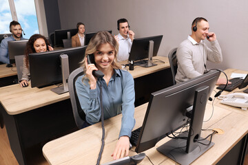 Wall Mural - Young call center operator talking on phone and her colleagues working in modern office