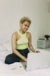 © shurkin_son - Young African female with lovely short blonde curls and nose ring having online consultation with her therapist, sitting with earphones on cozy white bed at her bedroom, smiling to laptop camera