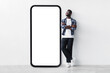 Full body length of black guy standing near huge giant smartphone with empty white screen template, mockup