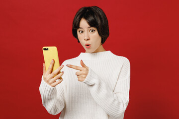 Young amazed astonished happy fun woman 20s wear white knitted sweater hold in hand use point index finger on mobile cell phone isolated on plain red color background studio. People lifestyle concept.