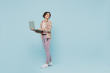 Wall Mural - Full body side view young smiling cheerful happy woman 20s wearing brown shirt hold use work on laptop pc computer look aside on area isolated on pastel plain light blue background studio portrait