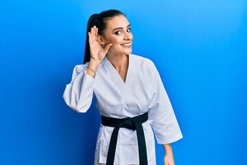 Wall Mural - Beautiful brunette young woman wearing karate fighter uniform with black belt smiling with hand over ear listening an hearing to rumor or gossip. deafness concept.