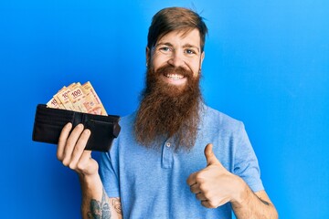 Sticker - Redhead man with long beard holding wallet with mexican pesos smiling happy and positive, thumb up doing excellent and approval sign